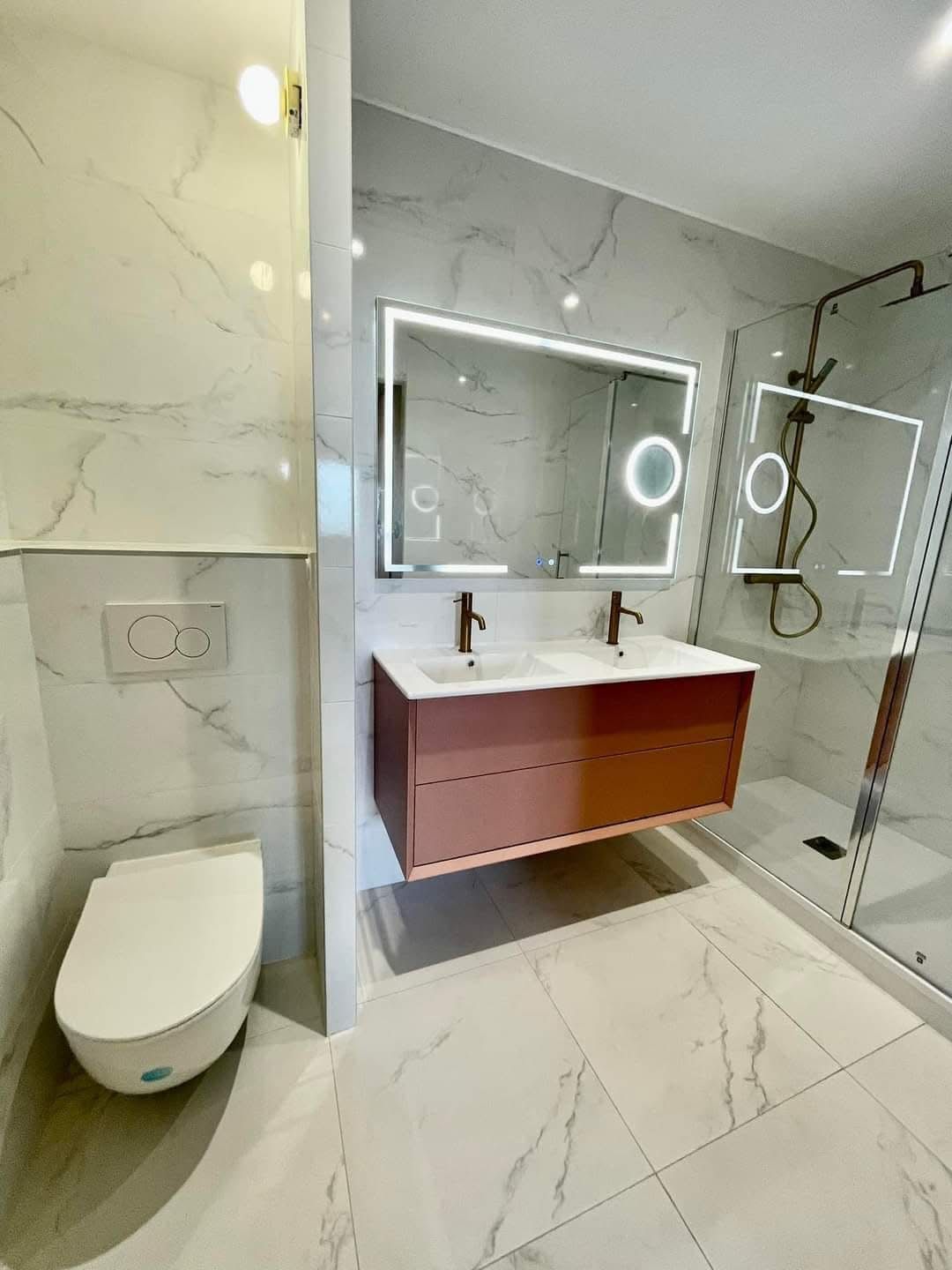Salle de bain en carrelage effet marbre avec double vasque sur meuble rose, miroir lumineux, WC suspendu et douche vitrée