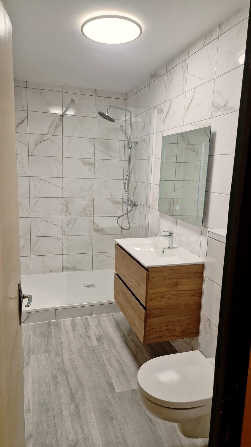 Salle de bain avec douche à paroi vitrée, meuble vasque en bois, miroir et WC suspendu